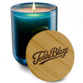 A lit scented candle in a blue-green glass jar with a wooden lid leaning in front, engraved with the brand name 'Teddy Blaze'.
