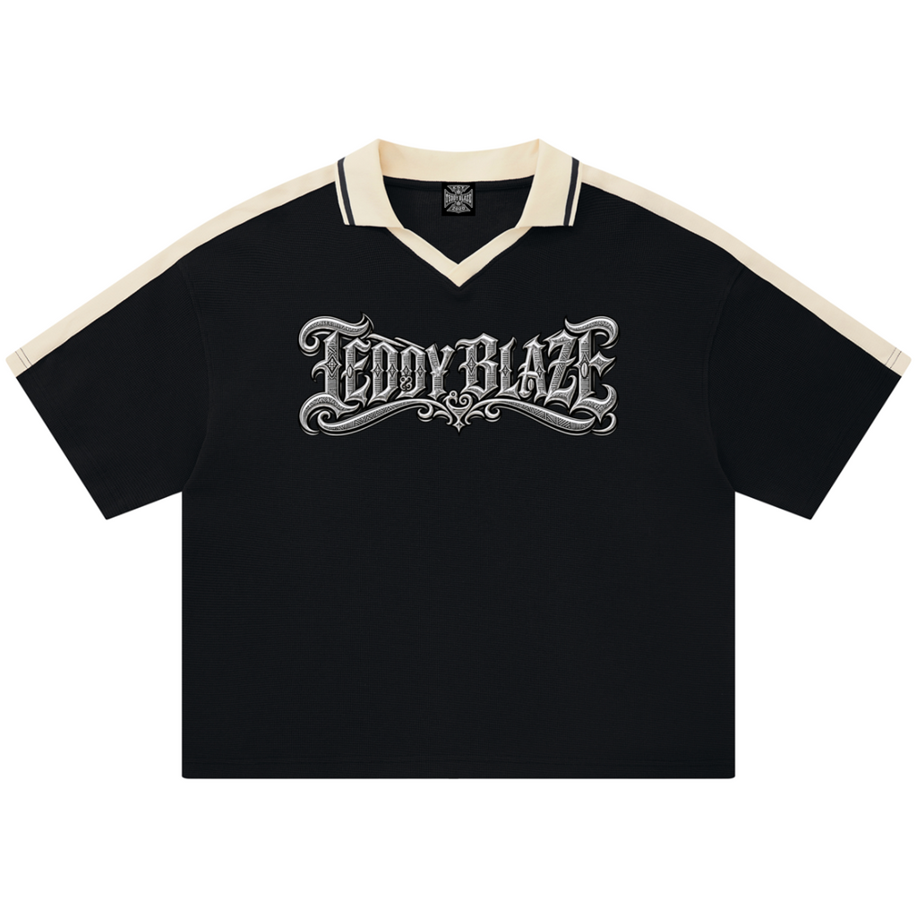 A black short-sleeve polo shirt with cream-colored shoulder panels and collar trim, featuring a large silver gothic 'TEDDY BLAZE' graphic across the chest, displayed on a clean white background.