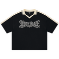 A black short-sleeve polo shirt with cream-colored shoulder panels and collar trim, featuring a large silver gothic 'TEDDY BLAZE' graphic across the chest, displayed on a clean white background.