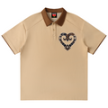 A beige short-sleeve polo shirt with a brown collar and sleeve trim, featuring a heart-shaped graphic on the left chest in a dark plaid pattern with a white star. The shirt is displayed flat against a solid white background.