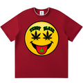 A red short-sleeve crewneck t-shirt featuring a large yellow smiley face graphic on the chest. The graphic has marijuana leaf eyes, the text 'TEDDY BLAZE' in black, and a red tongue with a green leaf detail.