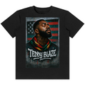 A black short-sleeve crewneck t-shirt featuring a large graphic print of a portrait of Teddy Blake wearing a red, green, and yellow scarf, set against a distressed American flag background with the text 'TEDDY BLAZE' and 'NEVER FORGET'.