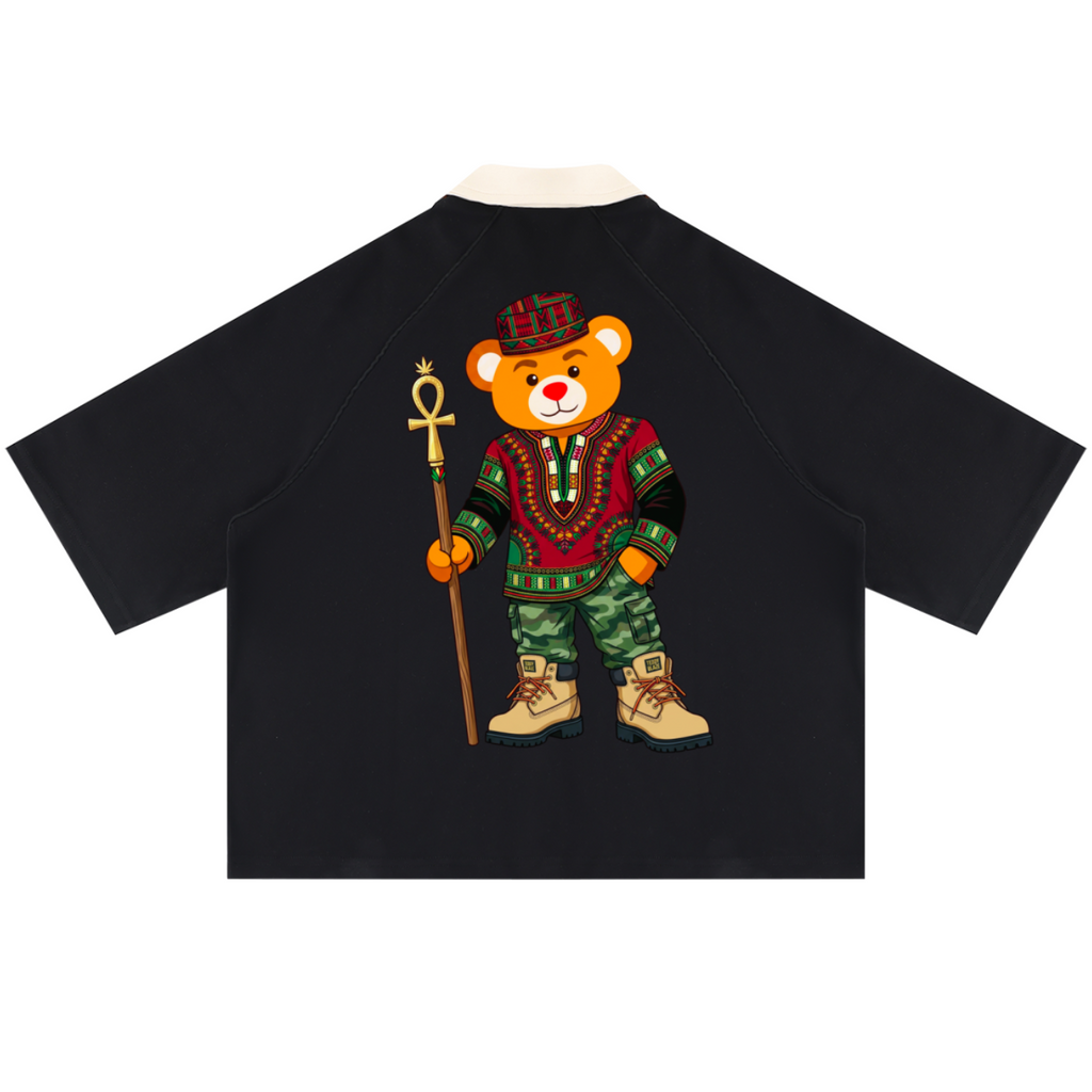 A black raglan-style short-sleeve shirt shown from the back, featuring a large graphic of a teddy bear dressed in colorful traditional African clothing and holding a staff, on a plain white background.