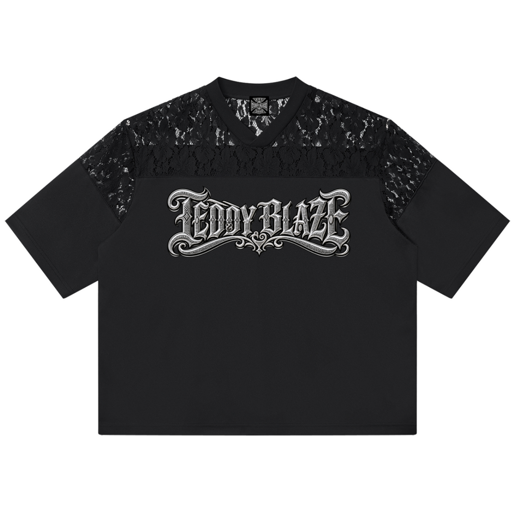 A black short-sleeve t-shirt with a sheer black lace yoke across the shoulders and upper chest, featuring a stylized silver 'TEDDY BLAZE' graphic printed across the chest, shown flat on a white background.