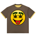 A brown ringer t-shirt with a large yellow smiley face graphic on the chest. The graphic features cannabis leaves for eyes, a black smile, and a red tongue with a green leaf.