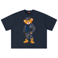 A navy blue short-sleeve t-shirt featuring a large graphic of a teddy bear wearing a matching plaid jacket, trousers, and a hat, with yellow boots. The t-shirt is displayed flat against a solid white background.
