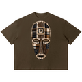 A dark brown short-sleeve t-shirt featuring a large, detailed graphic of an African tribal mask on the chest. The mask is constructed from various patterned fabrics in brown, beige, black, and blue, with intricate geometric designs.
