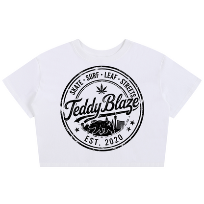 A white cropped short-sleeve t-shirt featuring a large black circular graphic on the front with the text 'Teddy Blaze' and 'EST. 2020' along with 'Skate Surf Leaf Street'.
