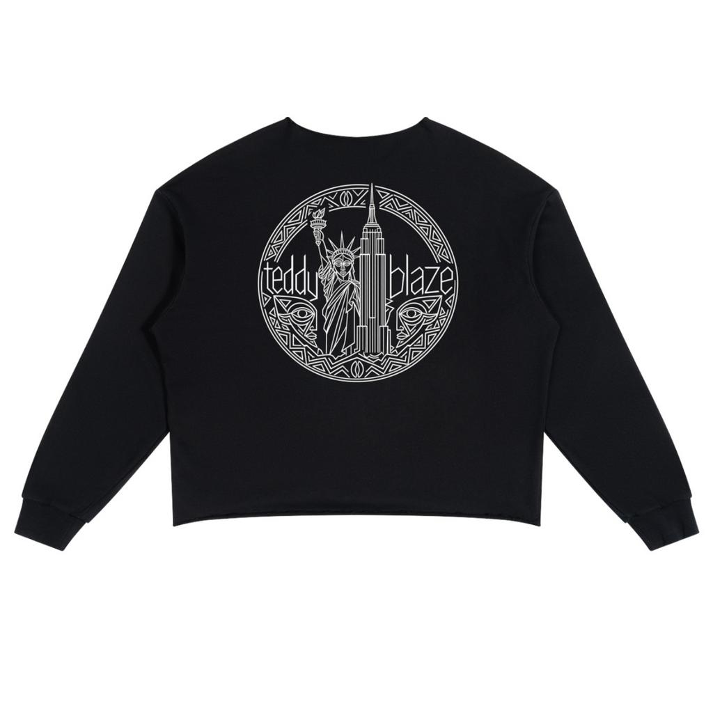 A black cropped sweatshirt with long sleeves, shown flat against a white background. The back of the sweatshirt features a large circular graphic in white, depicting the Statue of Liberty and the New York City skyline with the text 'teddy blaze'.