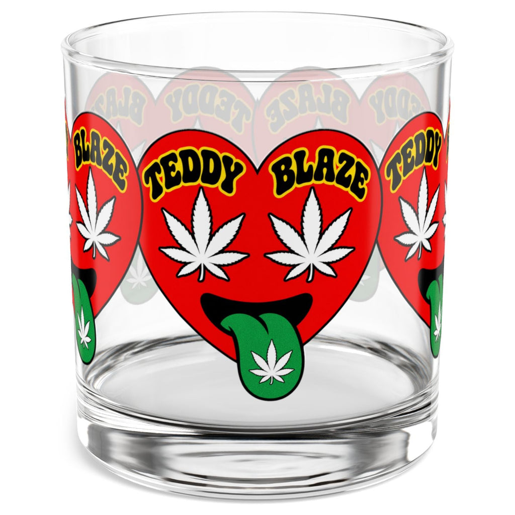 A clear glass rocks tumbler featuring a repeating red heart graphic with white cannabis leaves, green tongues, and yellow text reading 'TEDDY BLAZE'.
