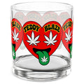 A clear glass rocks tumbler featuring a repeating red heart graphic with white cannabis leaves, green tongues, and yellow text reading 'TEDDY BLAZE'.