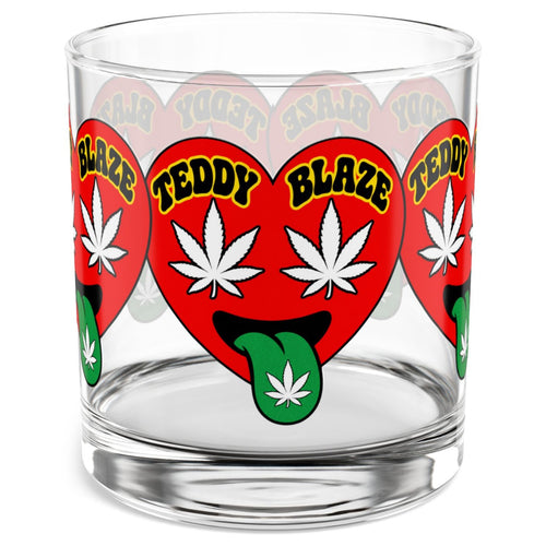 A clear glass rocks tumbler featuring a repeating red heart graphic with white cannabis leaves, green tongues, and yellow text reading 'TEDDY BLAZE'.