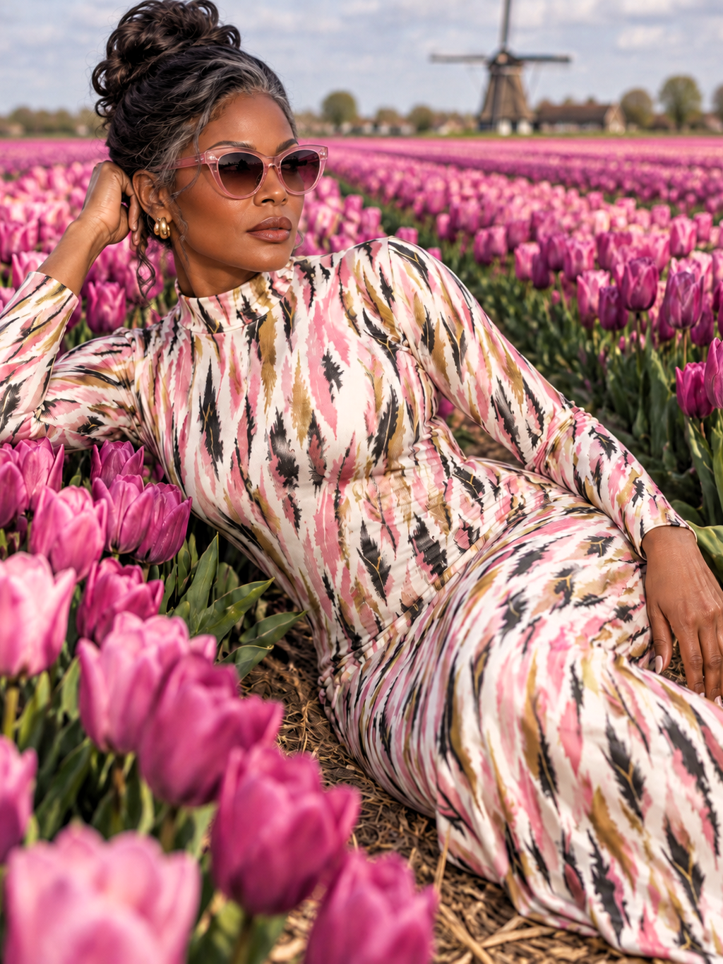 A woman reclining in a field of pink tulips, wearing a long-sleeve mock-neck dress with a pink, black, and beige ikat pattern. A windmill is visible in the background under a cloudy sky.