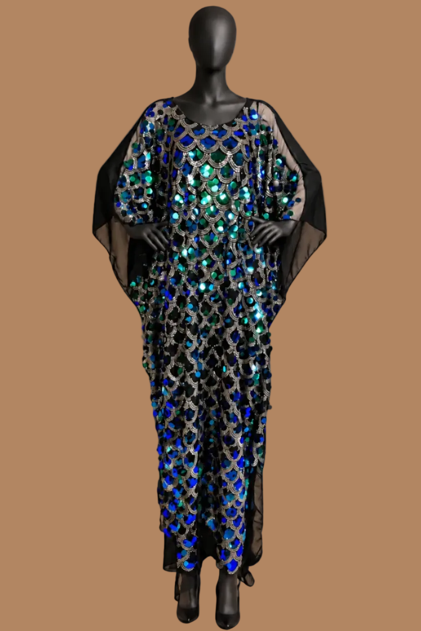 A floor-length black caftan-style dress with wide draped sleeves and an allover sequined geometric pattern, displayed on a black mannequin against a warm tan studio backdrop.