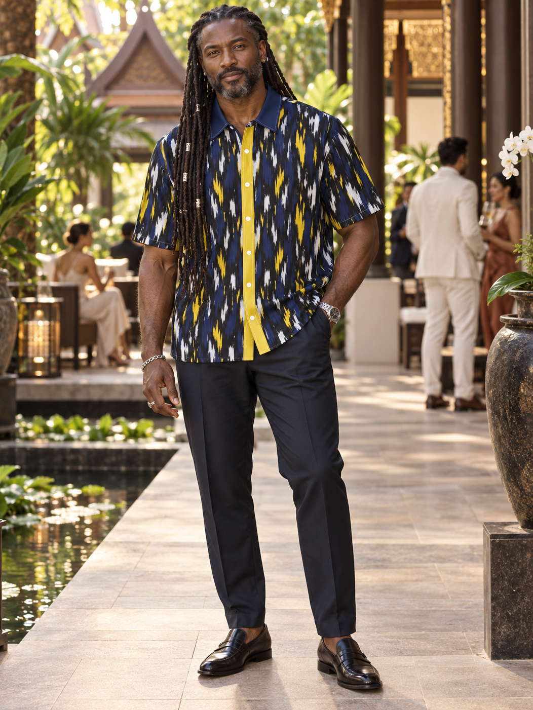 A man wearing a short-sleeve button-up shirt with a navy, yellow, and white ikat pattern, paired with navy trousers and black loafers, standing in a tropical resort courtyard.
