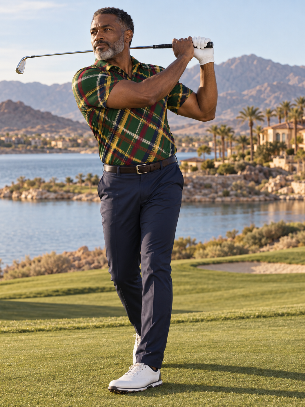A man wearing a green and red plaid short-sleeve polo shirt, navy pants, and white golf shoes is captured mid-swing on a golf course with water and mountains in the background.