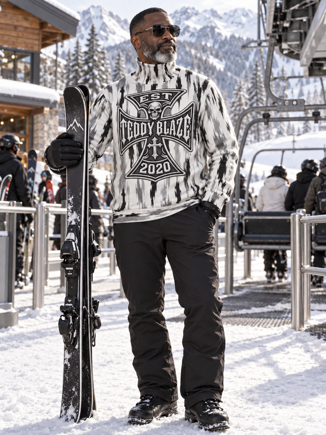 A man wearing a black and white tie-dye graphic sweatshirt with 'TEDDY BLAZE 2020' printed on the front, black snow pants, and black boots, standing in the snow at a ski resort and holding a snowboard.