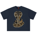 A navy blue short-sleeve t-shirt featuring a large, detailed graphic of a coiled cobra snake on the front. The snake is decorated with geometric patterns made of white dots, black and brown plaid, and gold accents.