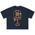 A navy blue short-sleeve t-shirt featuring a large, detailed graphic of a tribal-style mask on the chest. The mask is composed of beige, brown, and black plaid and geometric patterns.
