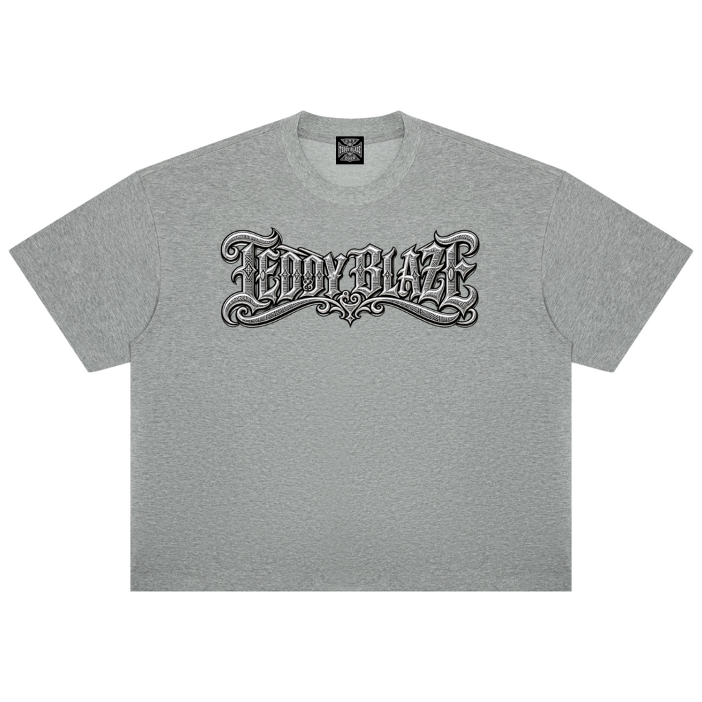 A heather gray short-sleeve t-shirt with a stylized black and white gothic-style 'TEDDY BLAZE' graphic printed across the chest, displayed flat against a solid white background.