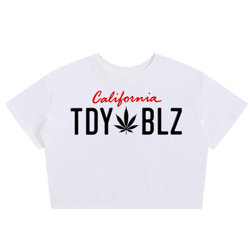 A white short-sleeve crop top t-shirt featuring a graphic print on the front. The design includes the word 'California' in red cursive script above the text 'TDY BLZ' in bold black letters, where the 'O' is replaced by a black cannabis leaf.