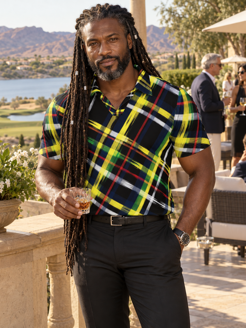 A man with long dreadlocks stands on an outdoor patio wearing a short-sleeve plaid button-up shirt with black trousers. The shirt has a bold plaid pattern in yellow, green, blue, red, and white over a dark base.