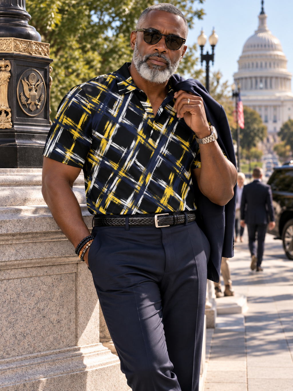 A man wearing a navy blue short-sleeve button-down shirt with a yellow and white plaid pattern, navy trousers, and sunglasses, leaning against a stone column outdoors with the US Capitol building in the background.