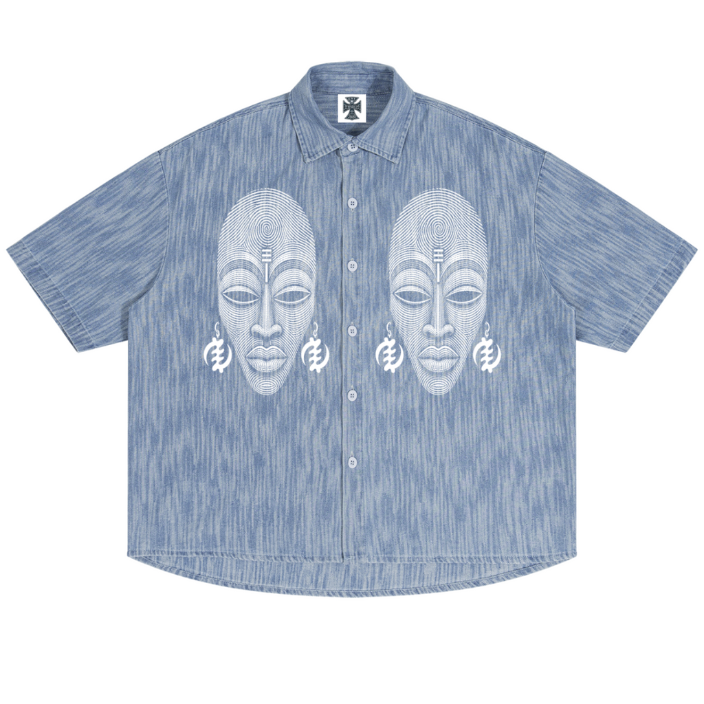 A short-sleeve button-up shirt in a heathered light blue fabric, featuring a white graphic print of two stylized African masks with ear ornaments across the chest.